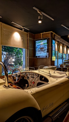 A classic, vintage car is parked indoors, featuring a polished white exterior and an open-top design. The car's dashboard is equipped with several dials and a wooden steering wheel. The interior environment is dimly lit with spotlights, while a large window offers a view of trees outside. There is also a framed picture of another car on the wall.