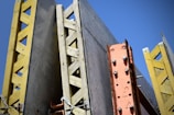 A structural framework composed of large, vertical concrete panels supported by wooden and metal beams. The beams are prominently yellow and orange, creating contrasting lines and textures against a clear blue sky.