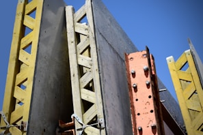 A structural framework composed of large, vertical concrete panels supported by wooden and metal beams. The beams are prominently yellow and orange, creating contrasting lines and textures against a clear blue sky.