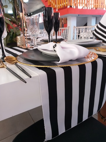 A sophisticated table setting features a black and white striped table runner with elegant black stemware, crystal glassware, and gold accents. Each place setting includes a gray plate topped with a white napkin carefully folded and held by a napkin ring. The background shows a modern, vibrant dining area with red and wooden elements.