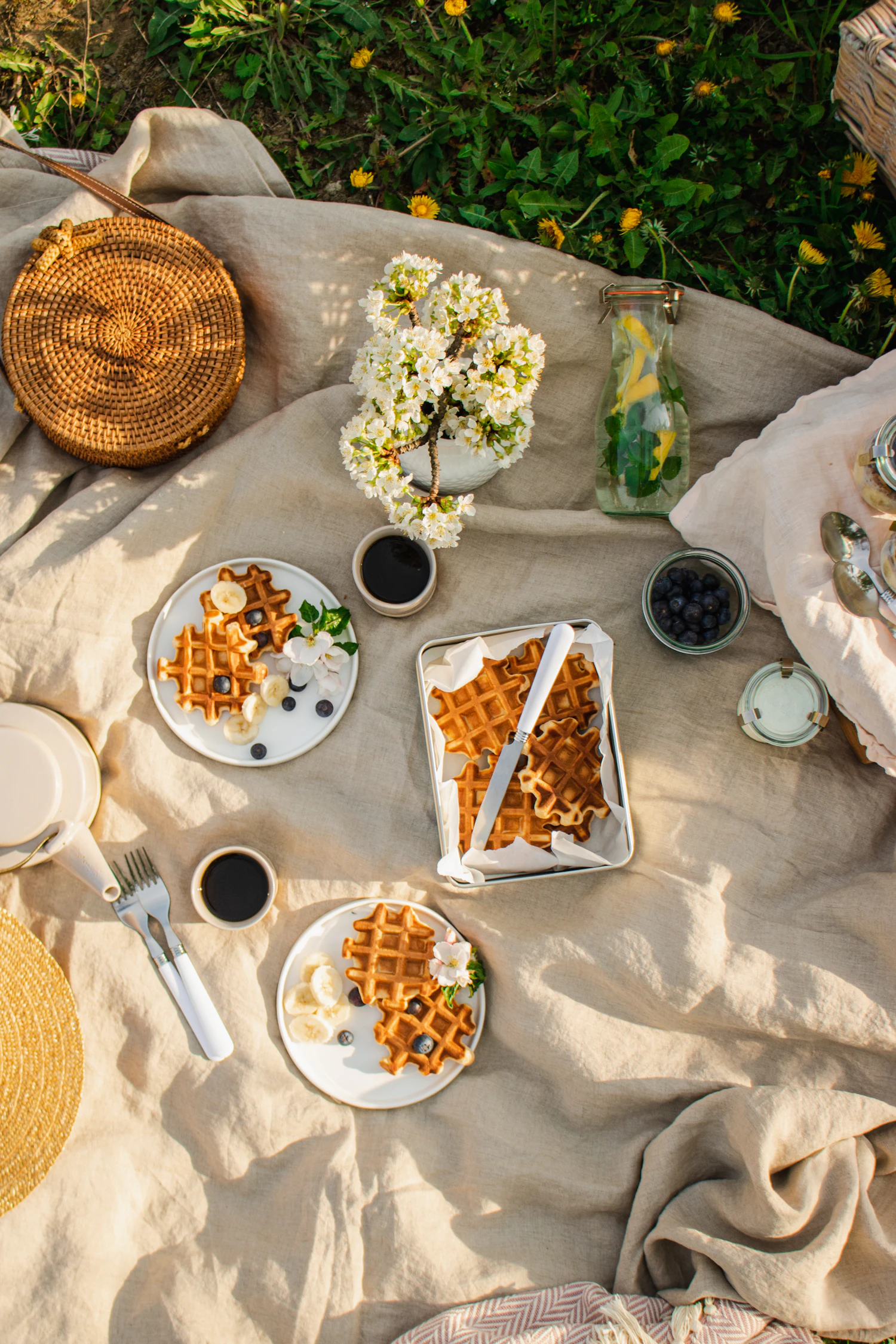 From above: plates of waffles, a dish of blueberries, coffee cups and white flowers on a beige linen blanket