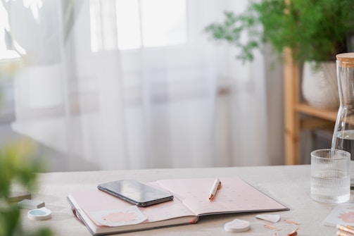A serene workspace featuring a table with a notebook, a pen, and a smartphone placed on top. The notebook lies open, and a glass of water with a cork stopper is nearby. In the background, there are blurred green plants and white curtains, allowing soft natural light to enter.