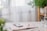 A serene workspace featuring a table with a notebook, a pen, and a smartphone placed on top. The notebook lies open, and a glass of water with a cork stopper is nearby. In the background, there are blurred green plants and white curtains, allowing soft natural light to enter.