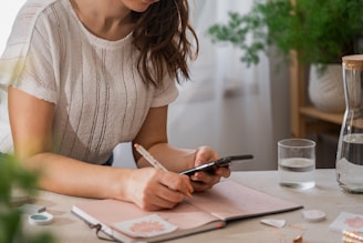 A person wearing a white knit short-sleeve top is holding a pen and a smartphone while leaning over a table. An open notebook with a floral design on the left page is on the table, alongside a glass of water and a carafe. There are green plants in the background, and soft, natural lighting fills the room.
