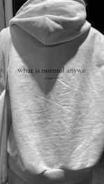 A person is wearing a hoodie with a text printed on the back. The text reads 'what is normal anyway' followed by 'YOUNG POETS'. The image is in black and white, and the hoodie appears to be light-colored.