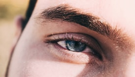 A close-up view of a person's eye featuring thick eyebrows and long lashes. The image highlights the intricate details of the skin texture and the natural eye color.