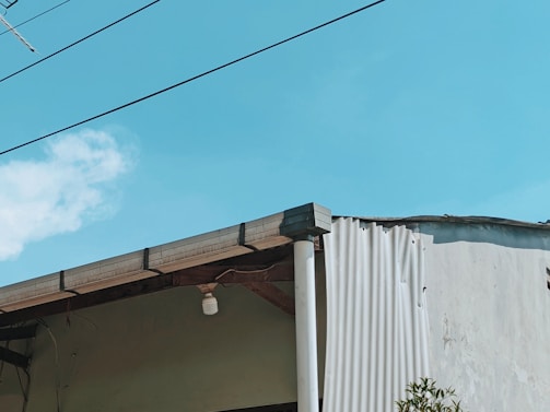 A portion of a building with a slanted roof is visible against a bright blue sky. The roof has a small, single light fixture hanging from it. There are wires running across the image, and part of the building's wall appears to be unfinished or damaged, showing corrugated sheeting. A small section of foliage is visible in the lower right corner.