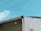 A portion of a building with a slanted roof is visible against a bright blue sky. The roof has a small, single light fixture hanging from it. There are wires running across the image, and part of the building's wall appears to be unfinished or damaged, showing corrugated sheeting. A small section of foliage is visible in the lower right corner.