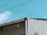 A portion of a building with a slanted roof is visible against a bright blue sky. The roof has a small, single light fixture hanging from it. There are wires running across the image, and part of the building's wall appears to be unfinished or damaged, showing corrugated sheeting. A small section of foliage is visible in the lower right corner.