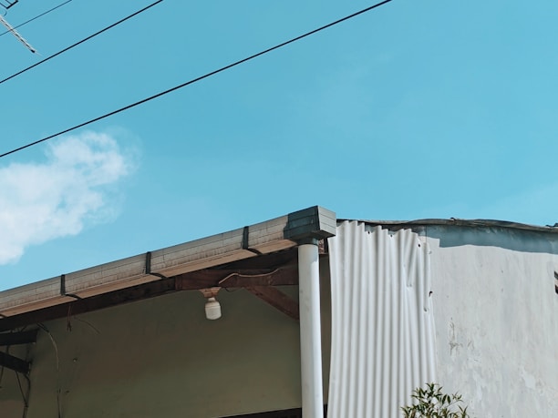 A portion of a building with a slanted roof is visible against a bright blue sky. The roof has a small, single light fixture hanging from it. There are wires running across the image, and part of the building's wall appears to be unfinished or damaged, showing corrugated sheeting. A small section of foliage is visible in the lower right corner.
