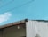 A portion of a building with a slanted roof is visible against a bright blue sky. The roof has a small, single light fixture hanging from it. There are wires running across the image, and part of the building's wall appears to be unfinished or damaged, showing corrugated sheeting. A small section of foliage is visible in the lower right corner.