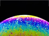 A vibrant, swirling mixture of colors with a domed shape, resembling a soap bubble under light. The colors include vivid yellows, purples, and blues, creating an abstract, marbled effect with a glossy texture. The background is a deep black, enhancing the luminous appearance of the colorful surface.