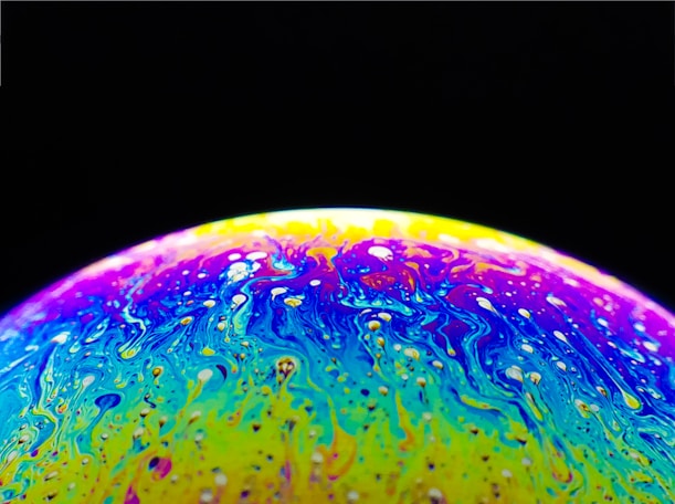 A vibrant, swirling mixture of colors with a domed shape, resembling a soap bubble under light. The colors include vivid yellows, purples, and blues, creating an abstract, marbled effect with a glossy texture. The background is a deep black, enhancing the luminous appearance of the colorful surface.