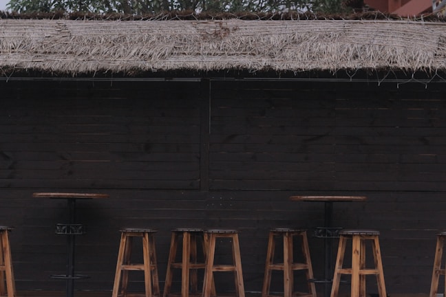 A rustic outdoor bar setup featuring a dark wooden counter with multiple wooden bar stools and tables. Above, a straw thatched roof is visible, providing a natural and earthy aesthetic. The area is devoid of people, giving it a serene and quiet atmosphere.