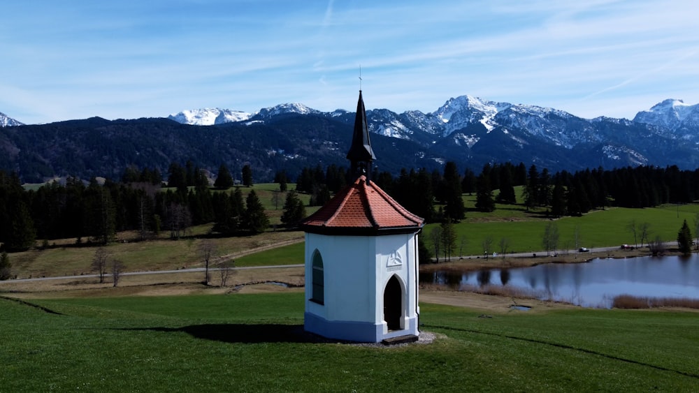 a small white building with a red roof by a lake with mountains in the background