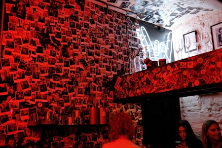 A vibrant room with walls completely covered in countless photographs, giving it a collage-like appearance. The lighting casts a red hue over the scene. Several people are present, two of whom appear to be using smartphones. Above a shelf adorned with stickers are empty wine glasses and bottles, with a neon peace sign in the background.