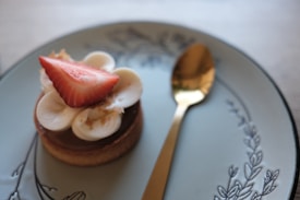 A small pastry topped with a dollop of cream and a slice of fresh strawberry sits elegantly on a decorative plate. Next to it, a gold spoon adds a touch of sophistication. The plate features subtle, intricate designs along its edges.