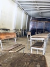 A room with wooden desks and benches neatly arranged in rows. The walls and ceiling are plain, with a simple structural design. A colorful striped fabric is visible in the background, possibly acting as a divider or curtain.