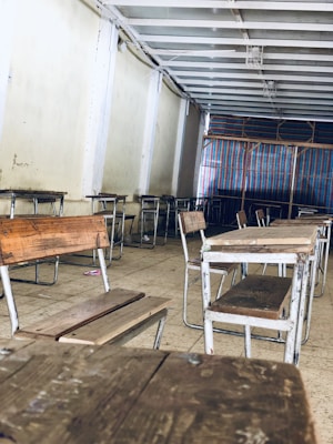 A room with wooden desks and benches neatly arranged in rows. The walls and ceiling are plain, with a simple structural design. A colorful striped fabric is visible in the background, possibly acting as a divider or curtain.