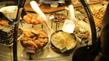 A variety of prepared foods are displayed behind a glass counter. Dishes include stuffed grape leaves, fried pastries, roast chicken, salads, and rice dishes. A person is serving rice into a takeout container while wearing gloves.
