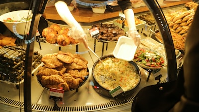 A variety of prepared foods are displayed behind a glass counter. Dishes include stuffed grape leaves, fried pastries, roast chicken, salads, and rice dishes. A person is serving rice into a takeout container while wearing gloves.