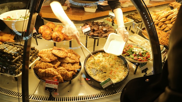 A variety of prepared foods are displayed behind a glass counter. Dishes include stuffed grape leaves, fried pastries, roast chicken, salads, and rice dishes. A person is serving rice into a takeout container while wearing gloves.
