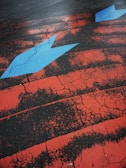 A cracked asphalt surface is painted with striking red lines and features a prominent blue arrow pointing to the right. The weathered texture of the paint contrasts sharply with the dark tarmac, creating a vivid and dynamic visual effect.