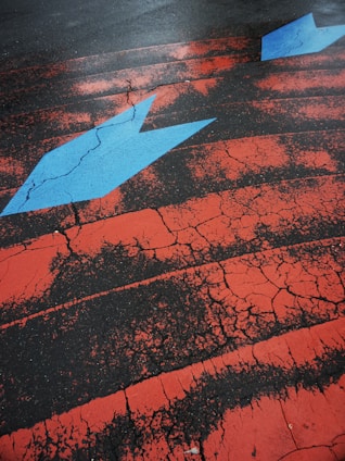 A cracked asphalt surface is painted with striking red lines and features a prominent blue arrow pointing to the right. The weathered texture of the paint contrasts sharply with the dark tarmac, creating a vivid and dynamic visual effect.