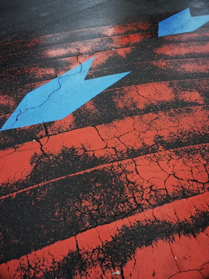 A cracked asphalt surface is painted with striking red lines and features a prominent blue arrow pointing to the right. The weathered texture of the paint contrasts sharply with the dark tarmac, creating a vivid and dynamic visual effect.