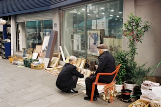 Two elderly individuals are examining items displayed on the ground in front of a store with glass windows. The items include various paintings, books, and possibly antique pieces. One person is sitting on an orange chair, while the other is squatting down. There are also potted plants and other miscellaneous items next to them.