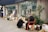Two elderly individuals are examining items displayed on the ground in front of a store with glass windows. The items include various paintings, books, and possibly antique pieces. One person is sitting on an orange chair, while the other is squatting down. There are also potted plants and other miscellaneous items next to them.