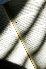An open book with text written in Greek is illuminated by sunlight, casting soft shadows across the pages. The text is neatly organized in columns with distinct chapter and verse numbers. The paper appears slightly aged, giving it a warm, yellowish tone.