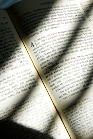 An open book with text written in Greek is illuminated by sunlight, casting soft shadows across the pages. The text is neatly organized in columns with distinct chapter and verse numbers. The paper appears slightly aged, giving it a warm, yellowish tone.
