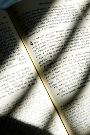 An open book with text written in Greek is illuminated by sunlight, casting soft shadows across the pages. The text is neatly organized in columns with distinct chapter and verse numbers. The paper appears slightly aged, giving it a warm, yellowish tone.