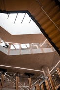 A modern architectural interior featuring multiple levels with glass railings and wooden accents. The open atrium space is illuminated by natural light coming through a large skylight. The design includes clean lines and minimalistic decor.
