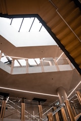 A modern architectural interior featuring multiple levels with glass railings and wooden accents. The open atrium space is illuminated by natural light coming through a large skylight. The design includes clean lines and minimalistic decor.