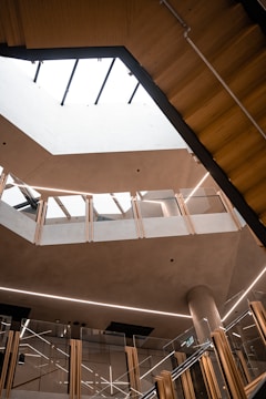 A modern architectural interior featuring multiple levels with glass railings and wooden accents. The open atrium space is illuminated by natural light coming through a large skylight. The design includes clean lines and minimalistic decor.