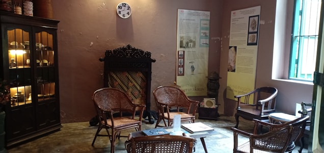 A cozy room furnished with several wicker chairs and a small table in the center, surrounded by informational posters on the walls. A wooden cabinet filled with small ceramics and artifacts is on the left, and a clock is mounted above a decorative fireplace on a warm-toned wall. The room is softly lit with natural light streaming through a window.