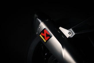 A close-up view of a sleek metallic exhaust pipe with a carbon fiber bracket, prominently featuring an Akrapovic sticker. The environment is dimly lit, emphasizing the polished metal and textured carbon fiber.