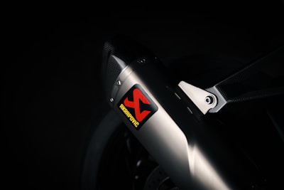 A close-up view of a sleek metallic exhaust pipe with a carbon fiber bracket, prominently featuring an Akrapovic sticker. The environment is dimly lit, emphasizing the polished metal and textured carbon fiber.