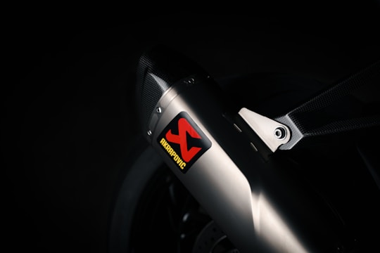 A close-up view of a sleek metallic exhaust pipe with a carbon fiber bracket, prominently featuring an Akrapovic sticker. The environment is dimly lit, emphasizing the polished metal and textured carbon fiber.