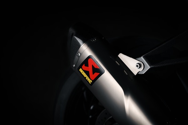 A close-up view of a sleek metallic exhaust pipe with a carbon fiber bracket, prominently featuring an Akrapovic sticker. The environment is dimly lit, emphasizing the polished metal and textured carbon fiber.