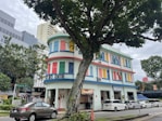 A vibrant, multi-story building with colorful window shutters in shades of red, blue, yellow, and green. It has a distinctive rounded corner and is labeled as an arts centre. The building is flanked by trees and is situated in an urban environment with modern office buildings nearby. Several cars are parked along the street, and a large tree partially obscures the view.