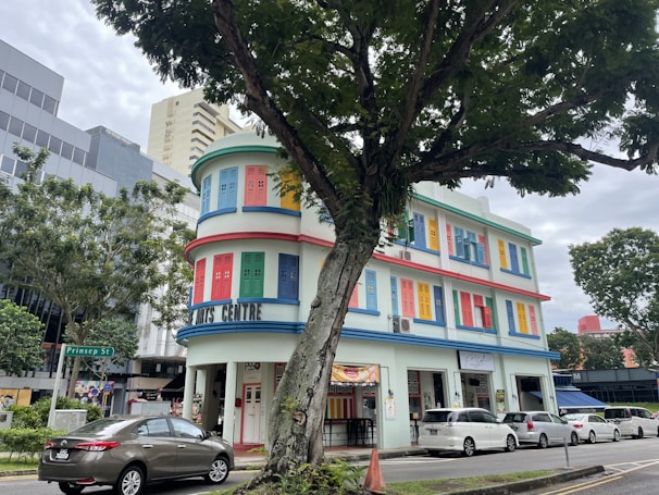 A vibrant, multi-story building with colorful window shutters in shades of red, blue, yellow, and green. It has a distinctive rounded corner and is labeled as an arts centre. The building is flanked by trees and is situated in an urban environment with modern office buildings nearby. Several cars are parked along the street, and a large tree partially obscures the view.