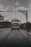 A tram labeled 'Metro do Porto' travels along railway tracks on a bridge, surrounded by architectural iron detailing. The scene has overcast skies, with people standing alongside the tracks, capturing a bustling urban environment.