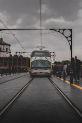 A tram labeled 'Metro do Porto' travels along railway tracks on a bridge, surrounded by architectural iron detailing. The scene has overcast skies, with people standing alongside the tracks, capturing a bustling urban environment.
