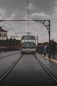 A tram labeled 'Metro do Porto' travels along railway tracks on a bridge, surrounded by architectural iron detailing. The scene has overcast skies, with people standing alongside the tracks, capturing a bustling urban environment.