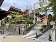 A traditional Japanese shrine set on a stone hilltop with stairs leading up. There are lush green trees and some red-leaved plants around the stone steps. Wooden structures with tiled roofs are visible, along with signs featuring Japanese characters.