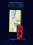 A gaming console with a screen displaying a video game scene, accompanied by two controllers, one colored red and the other blue. The devices are set against a dark backdrop, emphasizing the vibrant colors of the controllers.