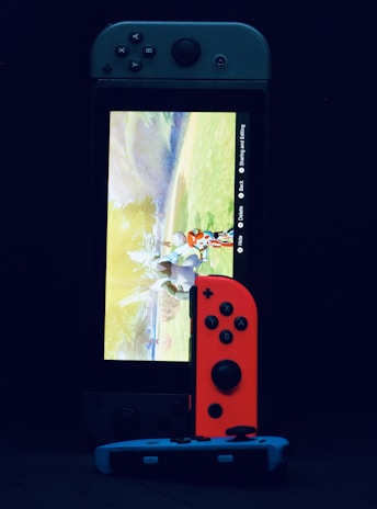 A gaming console with a screen displaying a video game scene, accompanied by two controllers, one colored red and the other blue. The devices are set against a dark backdrop, emphasizing the vibrant colors of the controllers.